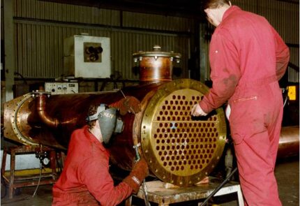 Two copper workers wear red boiler suits and soldering masks while soldering a copper still for distilling whisky